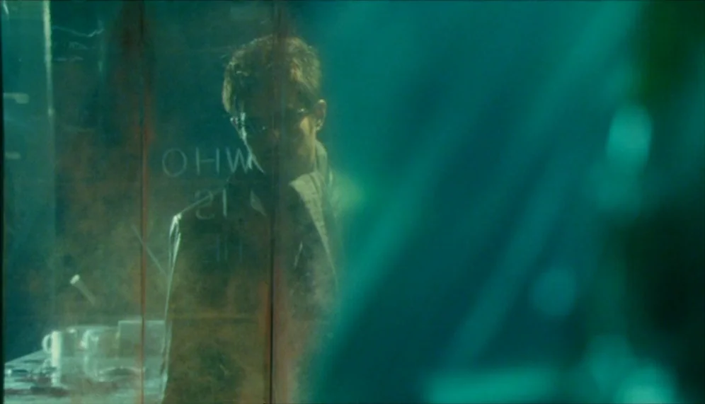 Gozu (2003) movie still: medium — A man stands behind a semi-transparent glass panel etched with the words WHO IS HE. The room is bathed in low-key…
