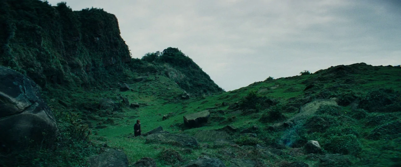 Silence (2016) movie still: wide — A green rocky hillside slopes upward toward a craggy peak under an overcast sky. A lone person stands at the base…