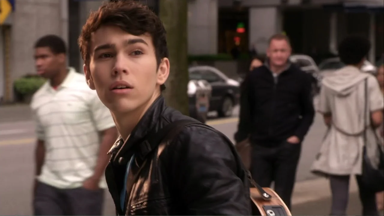 Rags (2012) movie still: medium — A bustling city sidewalk features blurred pedestrians and cars in the background. A young man with a guitar case…