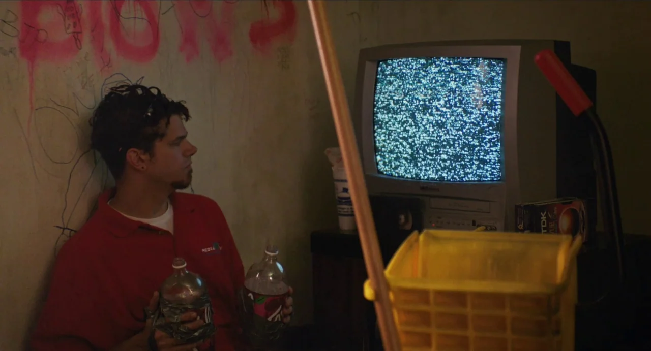 Relaxer (2018) movie still: medium — A dimly lit room features a plaster wall covered in faint, amateurish red spray paint. A man sits against the…