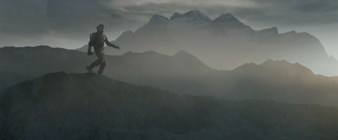 DISTANT (2026) movie still: wide — A desolate and craggy mountain landscape stretches under a cloudy and hazy sky. A person in a high-tech suit…