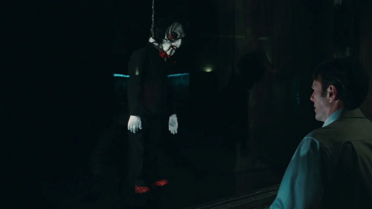 Saw VI movie still: medium — A dark observation room contains a glass partition separating a man from a puppet. The ventriloquist puppet hangs…