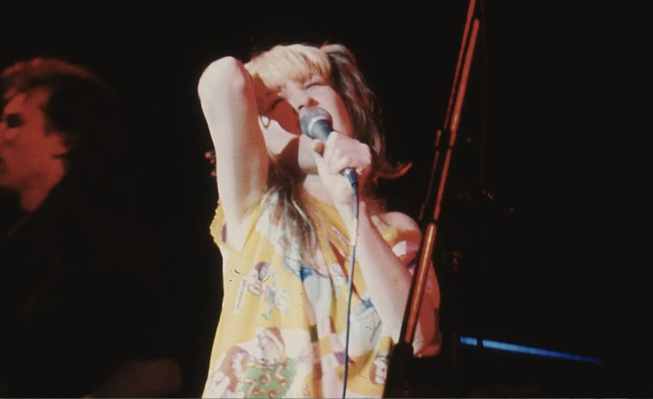 Let the Canary Sing (2023) movie still: medium — A dimly lit concert stage features a female singer positioned in front of a microphone. The singer,…