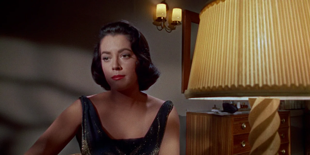 Imitation of Life (1959) movie still: medium — A dimly lit room contains a wooden dresser and wall-mounted lamp fixtures. A woman sits in the…