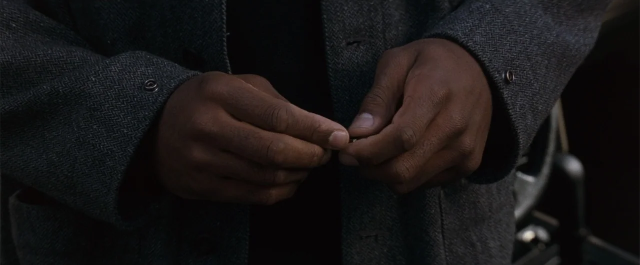 Fallen (2004) movie still: closeup — A close view shows dark-skinned hands manipulating a small metal item against a coat. The person wears a thick…