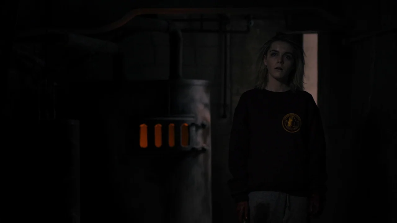 The Blackcoat’s Daughter (2015) movie still: medium — A dimly lit industrial basement features an old metal furnace with glowing vents. A young…