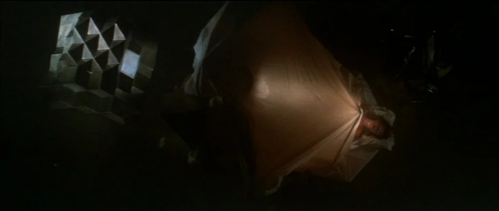 Demon Seed (1977) movie still: wide — A dark, void-like space contains a bed with a person lying under white sheets. The subject holds an internal…