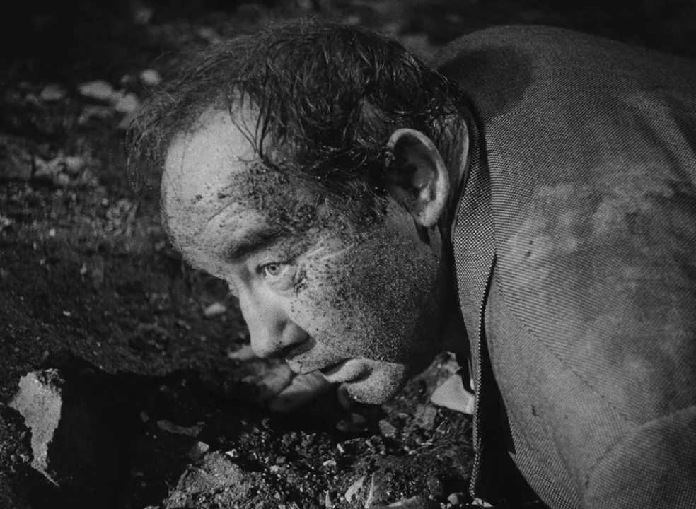 Il Bidone (1955) movie still: closeup — A dark, gritty outdoor setting features a man lying flat on dirt and gravel. The man's face and clothes are…