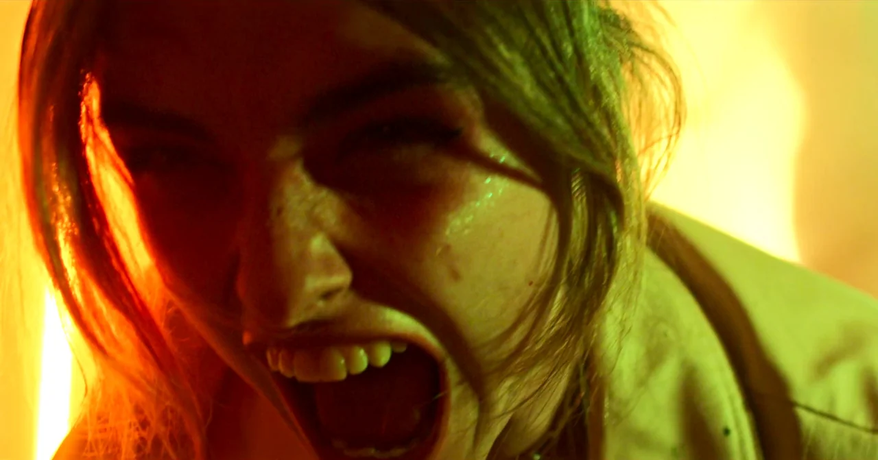 T Blockers (2023) movie still: closeup — A dark space lit by high-intensity red and yellow colored light sources. A young woman screams with her…