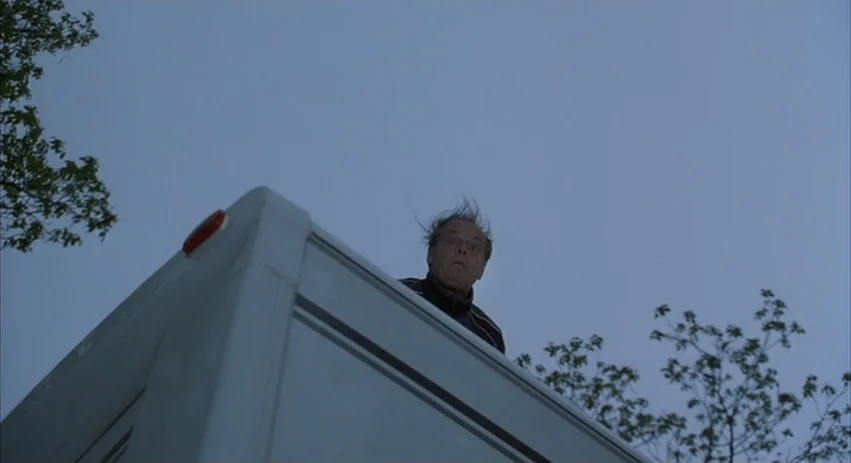 About Schmidt movie still: medium — A man stands atop a white vehicle roof against a clear, pale blue sky. The scene is shot from a low angle,…