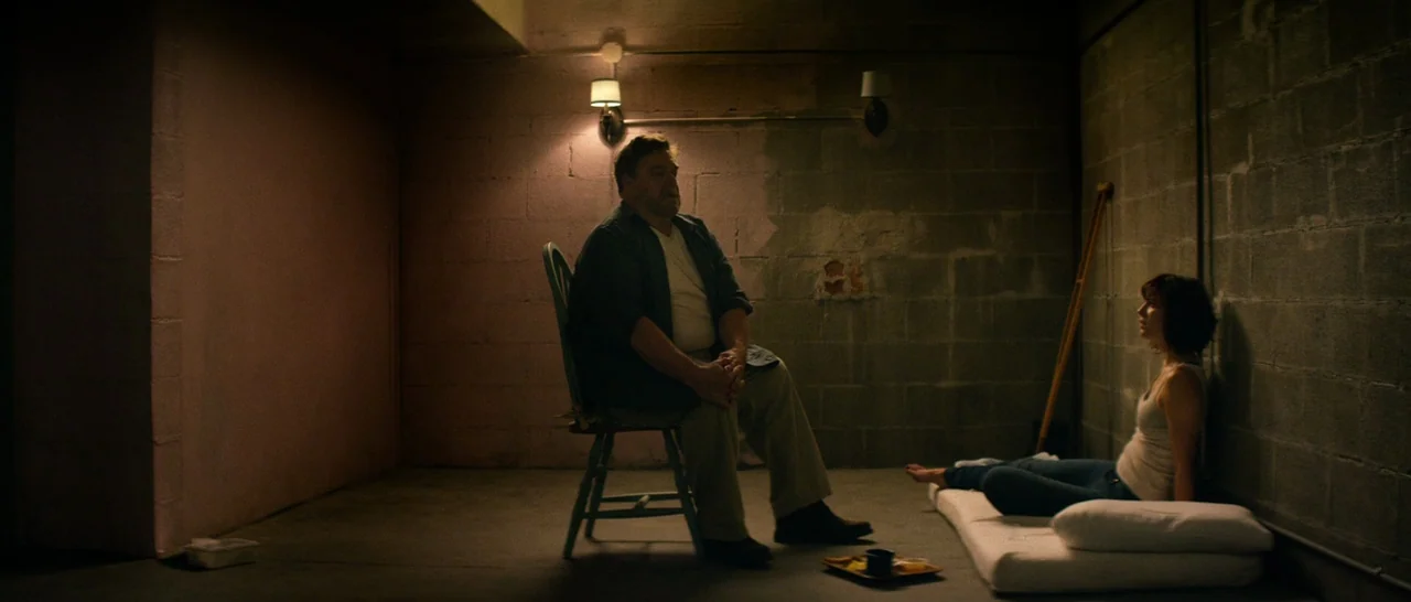 10 Cloverfield Lane (2016) movie still: wide — A basement room with bare cinder block walls and concrete floor. An older man sits on a wooden chair…