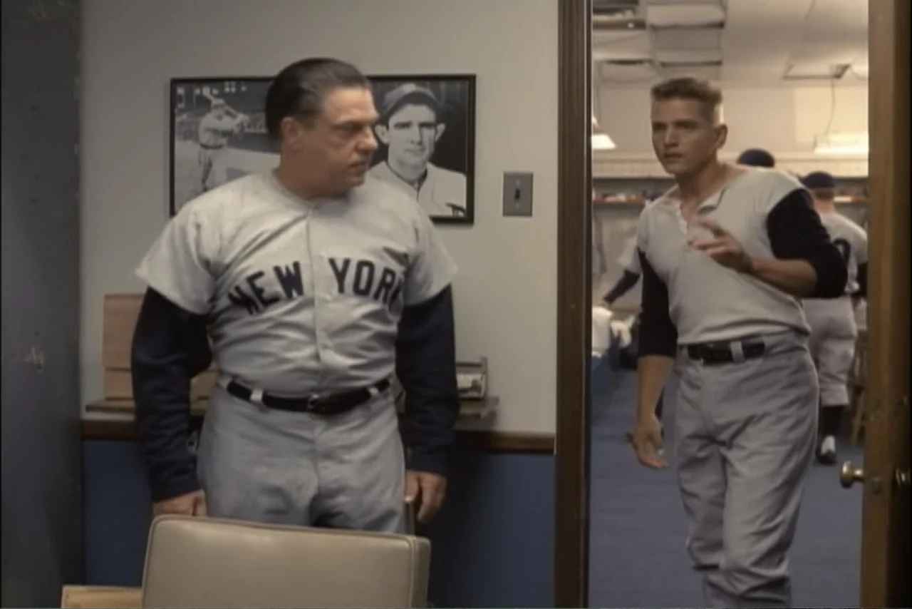 Unification of Japan 61 (2024) movie still: medium — A 1950s sports locker room features framed baseball photos on light-colored walls. Two players…