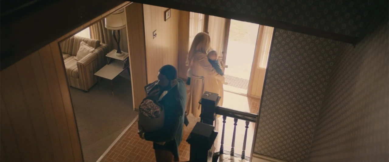 I'm Your Woman (2020) movie still: wide — A 1970s suburban house entry features wood paneling and vintage wallpaper. A man with groceries walks past…