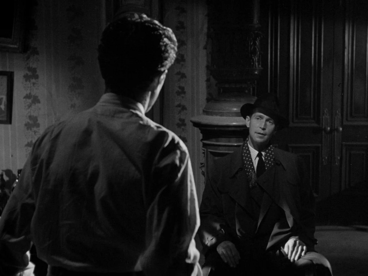 Phantom Lady (1944) movie still: medium — A dimly lit room with ornate wooden doors and patterned wallpaper. A man in a dark coat sits while talking…