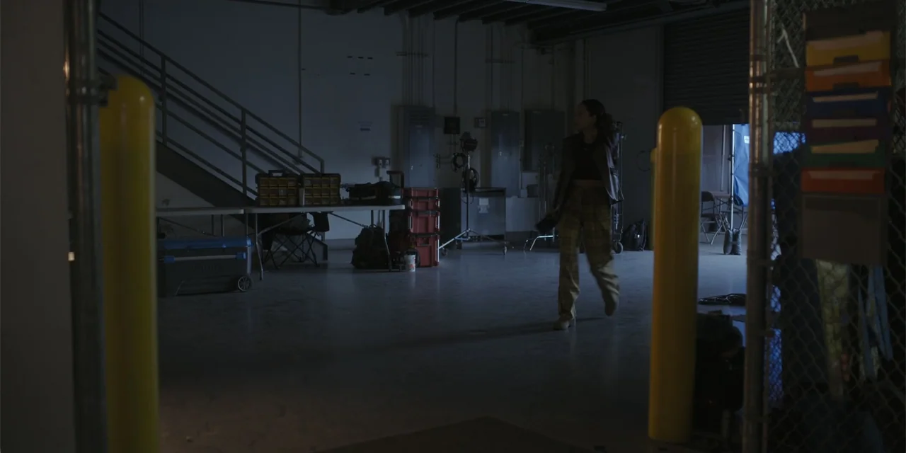 A Hollywood Christmas (2022) movie still: fullbody — A dimly lit industrial interior has a staircase and scattered equipment tables on the left. A…