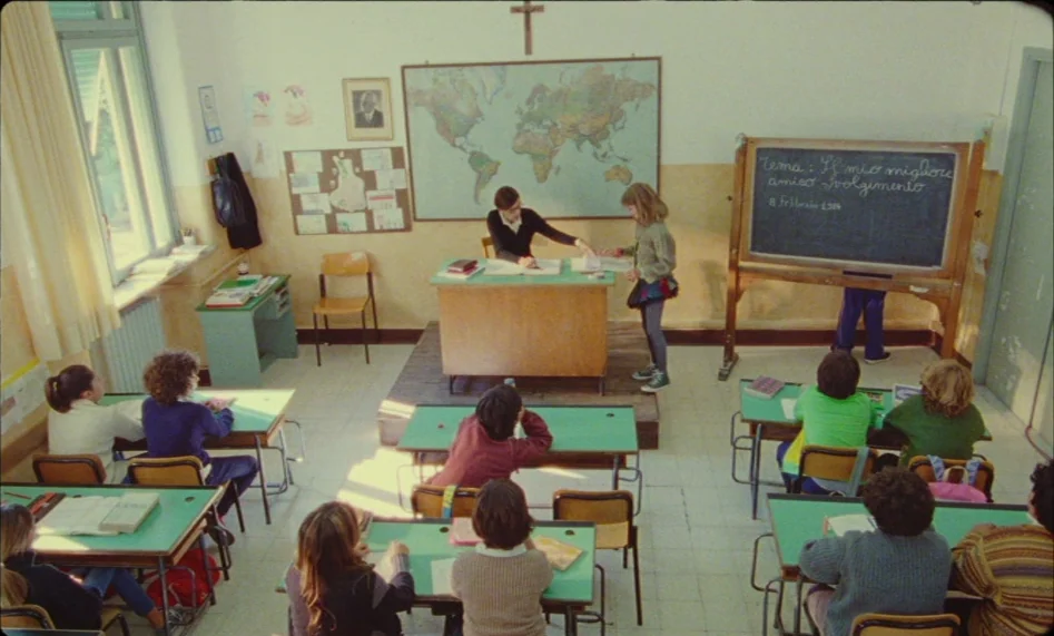 Misunderstood (2014) movie still: wide — A primary school room has rows of green-topped wooden desks and a wall map. A student stands near the…