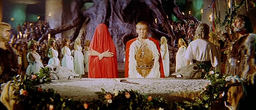 Erik The Conqueror (1961) movie still: wide — A stone altar adorned with flowers and animal skulls sits in a cavernous chamber. A man and a figure…