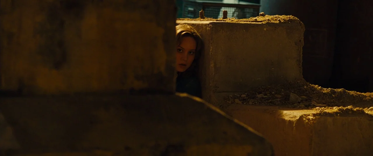 Free Fire (2016) movie still: medium — Large concrete blocks are stacked in a dark and dusty construction site at night. A young woman peeks around…