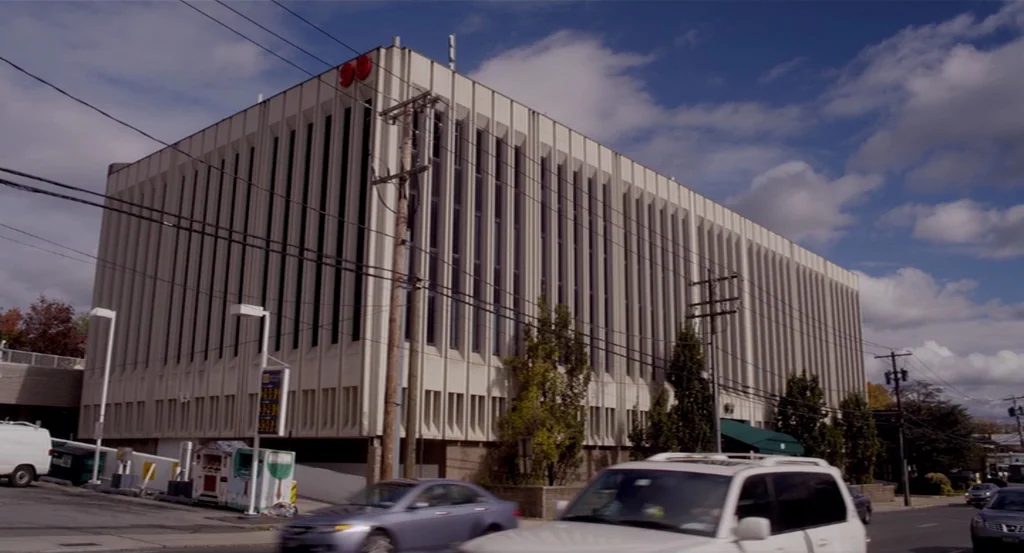 Dark Horse (2011) movie still: establishing — A tall, rectangular concrete office building with rhythmic vertical windows stands under a blue sky.…