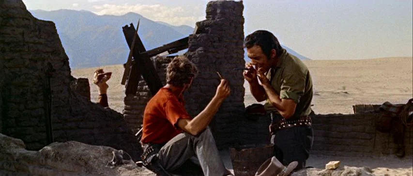 Ride Lonesome (1959) movie still: medium — A crumbling adobe structure sits amidst a desolate desert landscape with distant blue mountains. Two men…