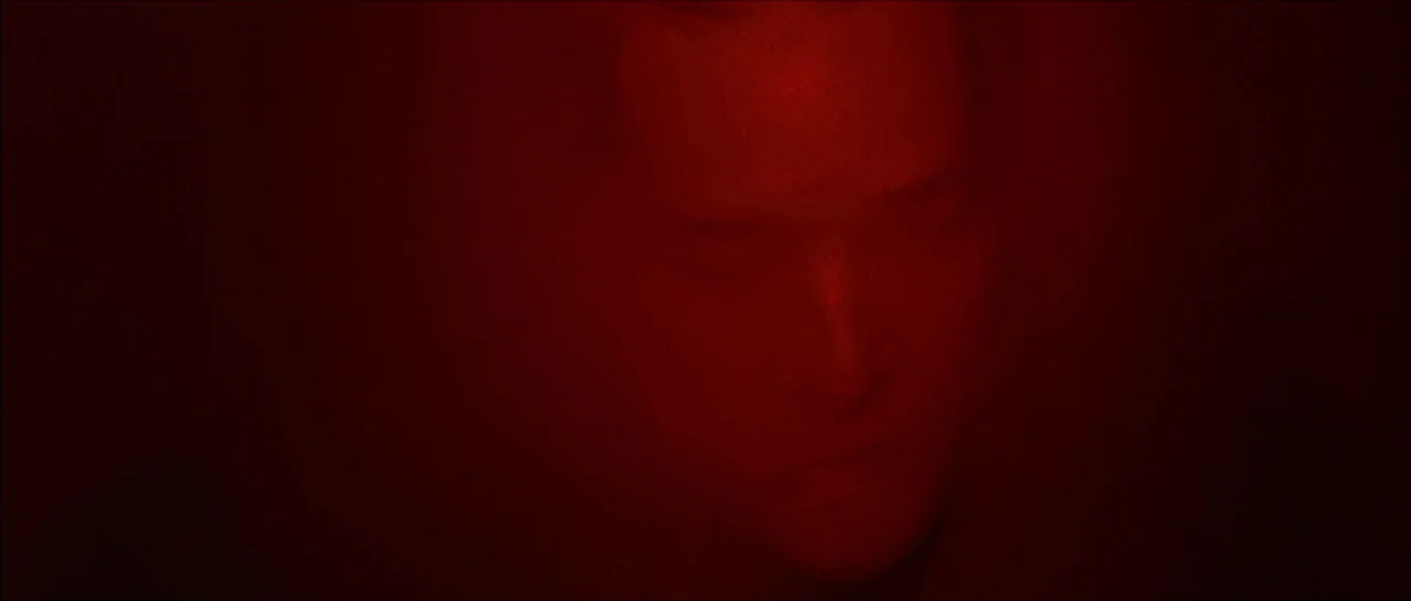 Fear X (2003) movie still: closeup — A dimly lit interior space is completely overwhelmed by a dense red light. A person with eyes closed is visible…