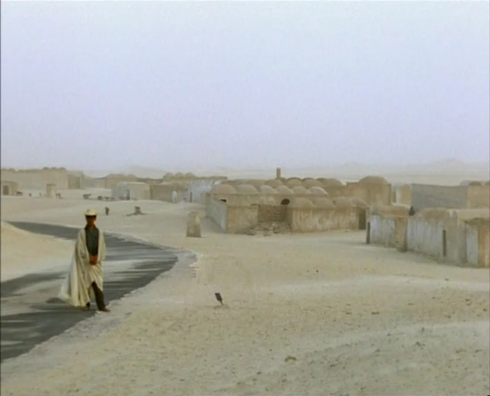 Fata Morgana (1971) movie still: wide — A remote desert settlement features low-slung, beige, domed structures along a dusty landscape. A man in…