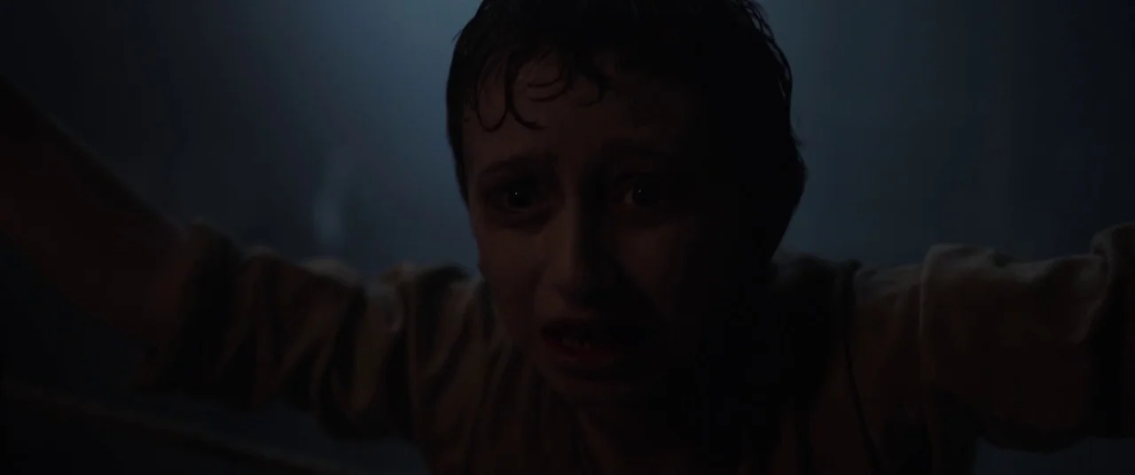 The Nun (2018) movie still: closeup — A dark, indistinct indoor space surrounds a person emerging from shadow. A young boy with wet hair and a plain…