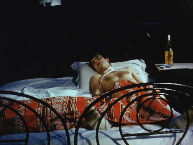 Our Lady of the Turks (1968) movie still: medium — A dimly lit bedroom features a dark metal bed frame and a patterned blanket. A man lies in the…