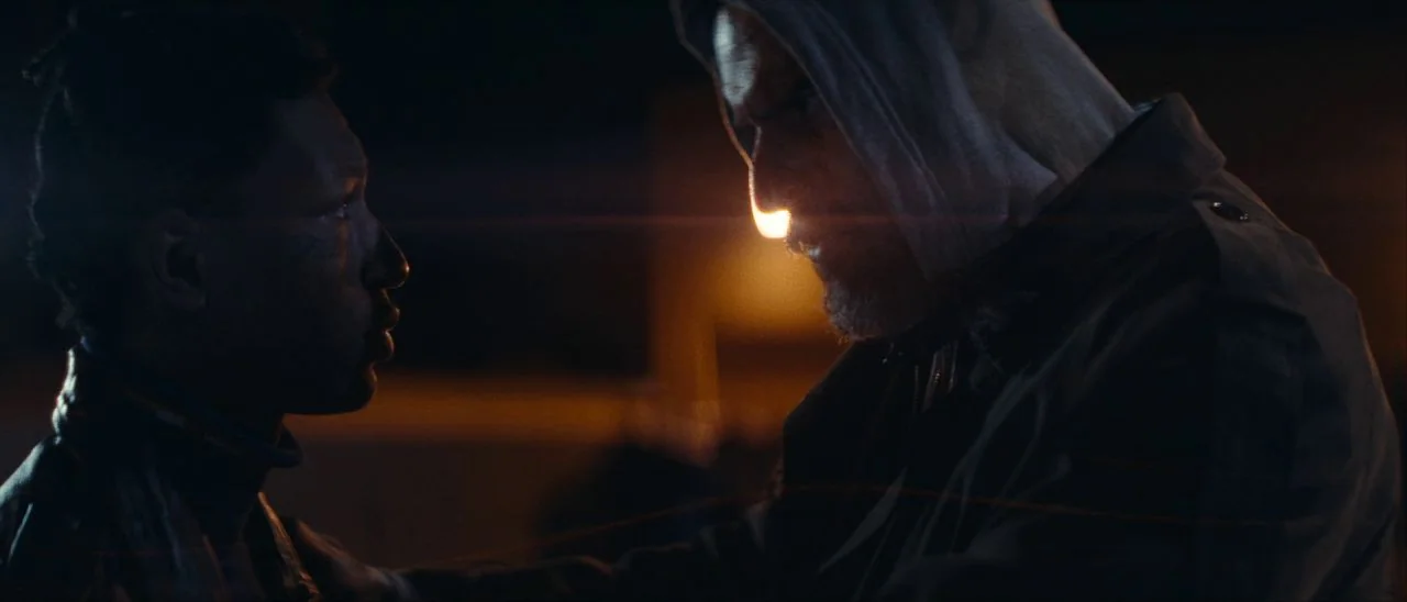 Archenemy (2020) movie still: medium — An outdoor city street at night illuminated by a single harsh, warm light source. An older man in a grey hood…