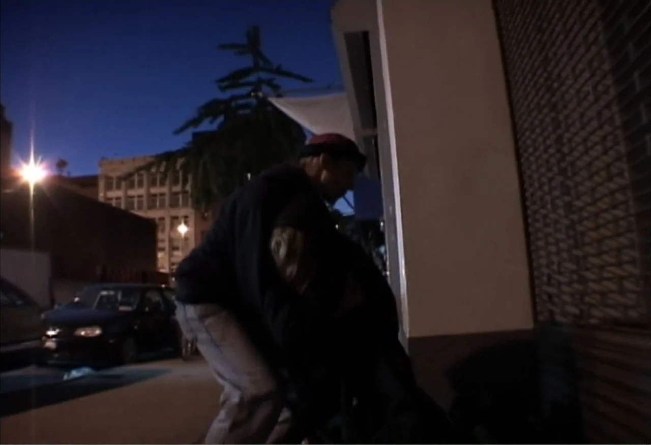 Dope Sick Love (2005) movie still: fullbody — A city street at night features a dark building wall and a parked car. A man in a dark sweatshirt and…