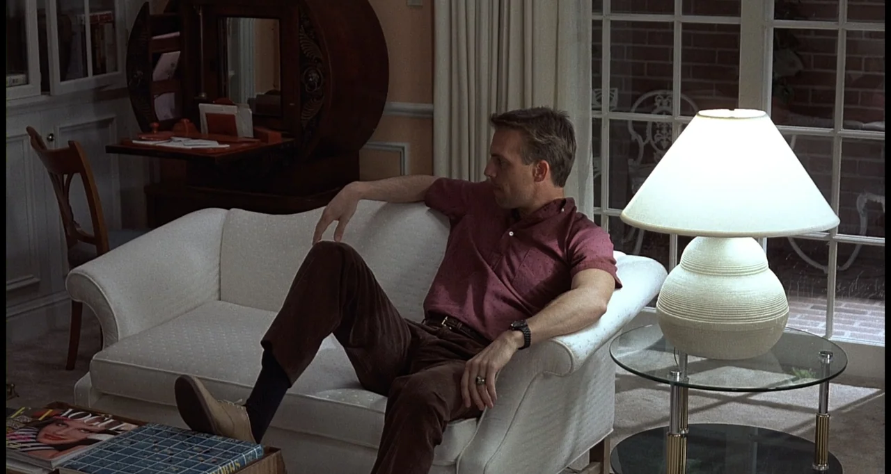 No Way Out (1987) movie still: medium — A well-lit living room contains a white couch, a lamp, and a small table. A man sits on the couch looking…