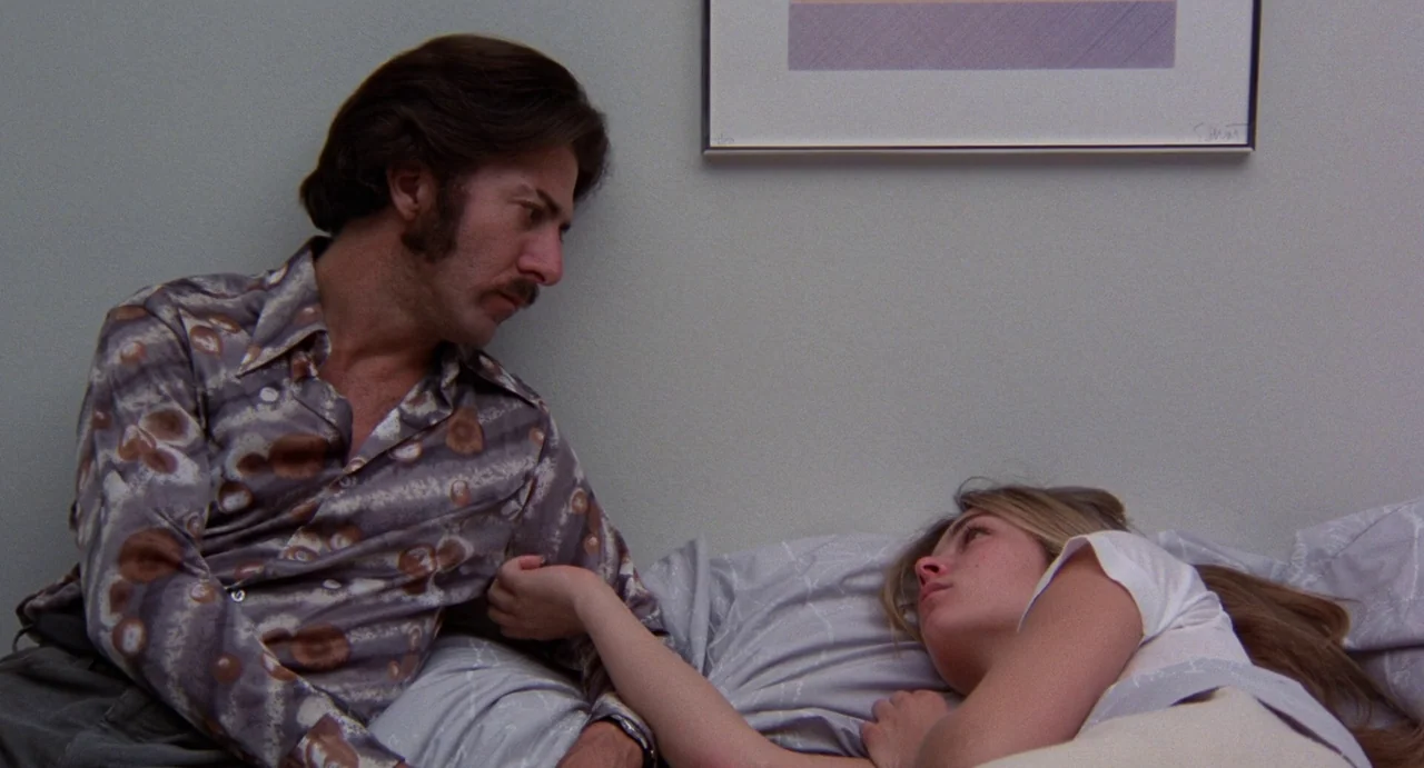 Straight Time (1978) movie still: medium — A man sits beside a woman who is lying on a bed. The wall features a simple framed abstract print.