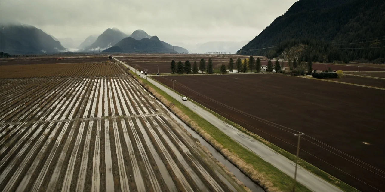 Playdate (2025) movie still: wide — An expansive valley floor features dark brown tilled fields and flooded planting beds. A single vehicle travels…