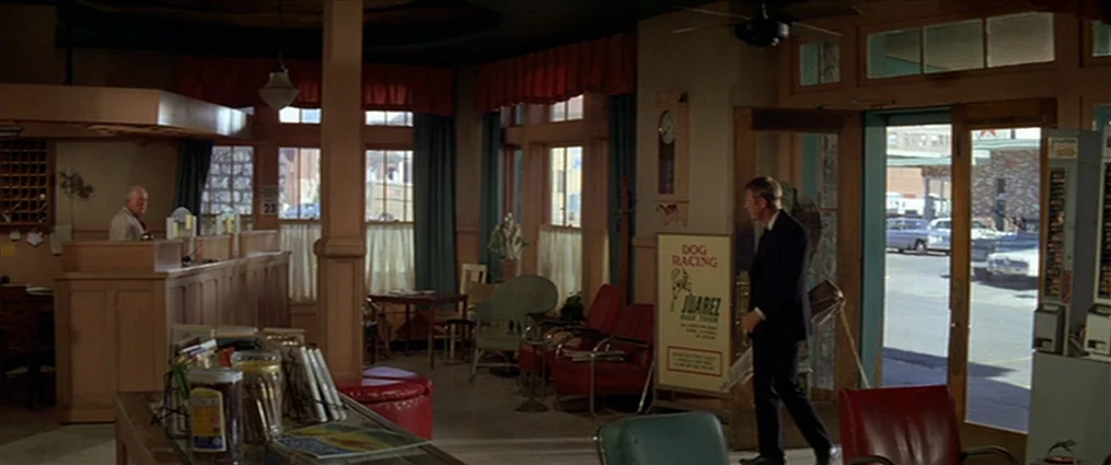 The Getaway (1972) movie still: wide — A dated hotel lobby features wooden counters, red chairs, and large glass entry doors. A man in a suit walks…