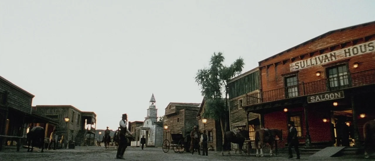 Renegade (2004) movie still: wide — A dusty main street in a classic wooden western town with a prominent saloon. Several men stand around the dirt…