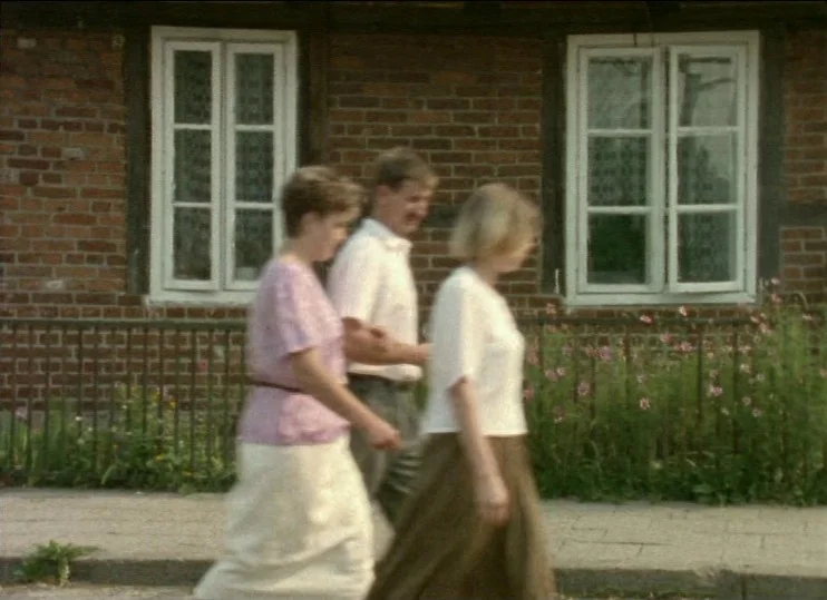 From The East (1993) movie still: fullbody — A red brick wall with two windows serves as a background for a sidewalk. Three people walk past the…