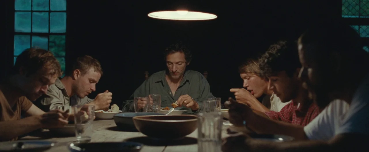 Martha Marcy May Marlene (2011) movie still: wide — A group of men sit at a long wooden table in a dimly lit room. They are eating dinner together…