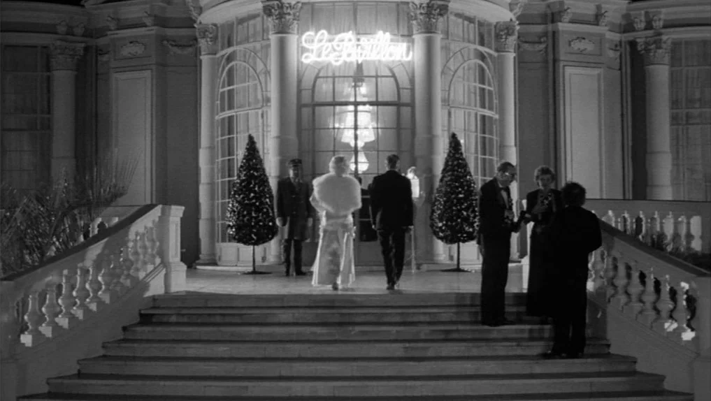 Under The Cherry Moon (1986) movie still: wide — A stone mansion entrance features a neon sign reading Le Pavillon above the doors. Several people…