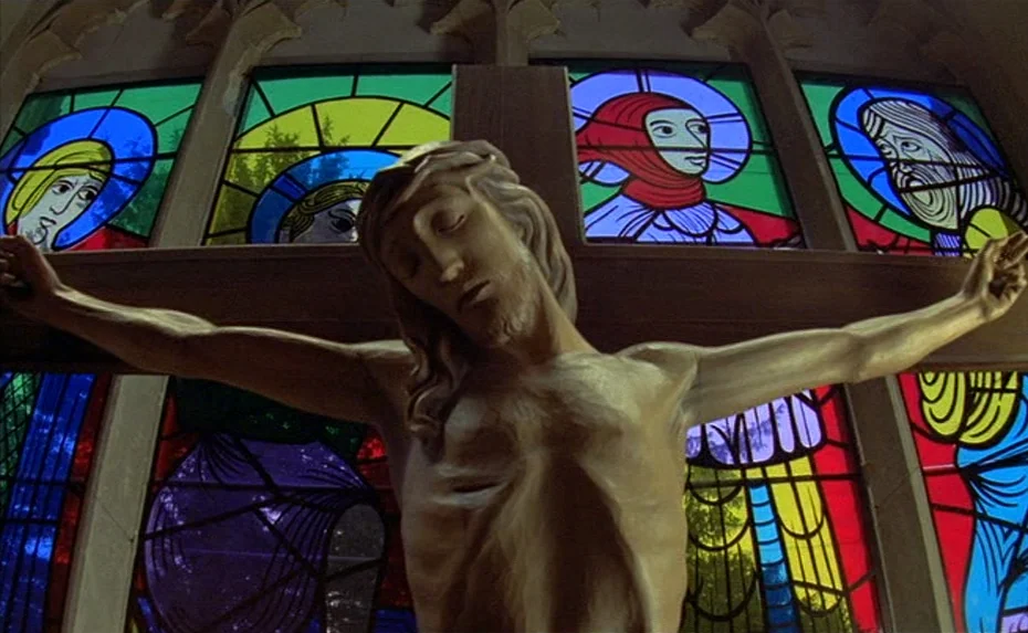 To The Devil a Daughter (1976) movie still: medium — A wooden crucifix stands before a multi-pane stained glass window in a church. The figure of…