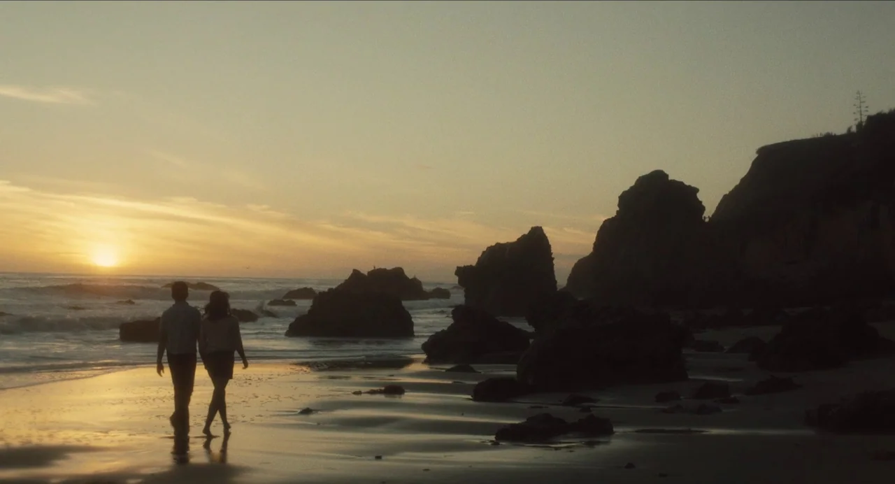Born To Be Blue (2015) movie still: wide — A rocky beach shoreline at sunset with dark jagged rocks protruding from the water. A man and woman hold…