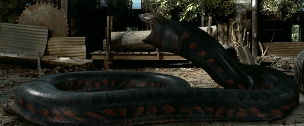 Anaconda (1997) movie still: medium — A cluttered, rustic industrial yard contains a large pile of logs and circular saws. A massive dark serpent…