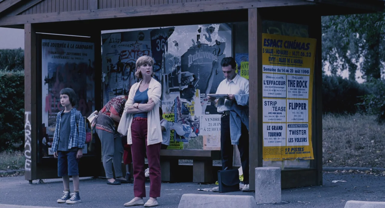 Ma Vie En Rose (1997) movie still: fullbody — A wooden bus shelter features posters for 1996 films like The Rock and Twister. A woman stands in the…