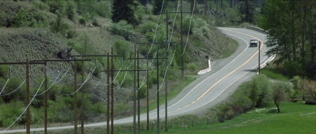 The Pledge (2001) movie still: wide — A winding asphalt road cuts through a green hillside with utility poles lining the shoulder. A single…