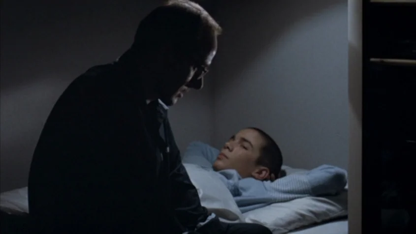 Benny’s Video (1992) movie still: medium — A dimly lit room with a man sitting beside a young boy lying down. The boy rests on a bed while the man…