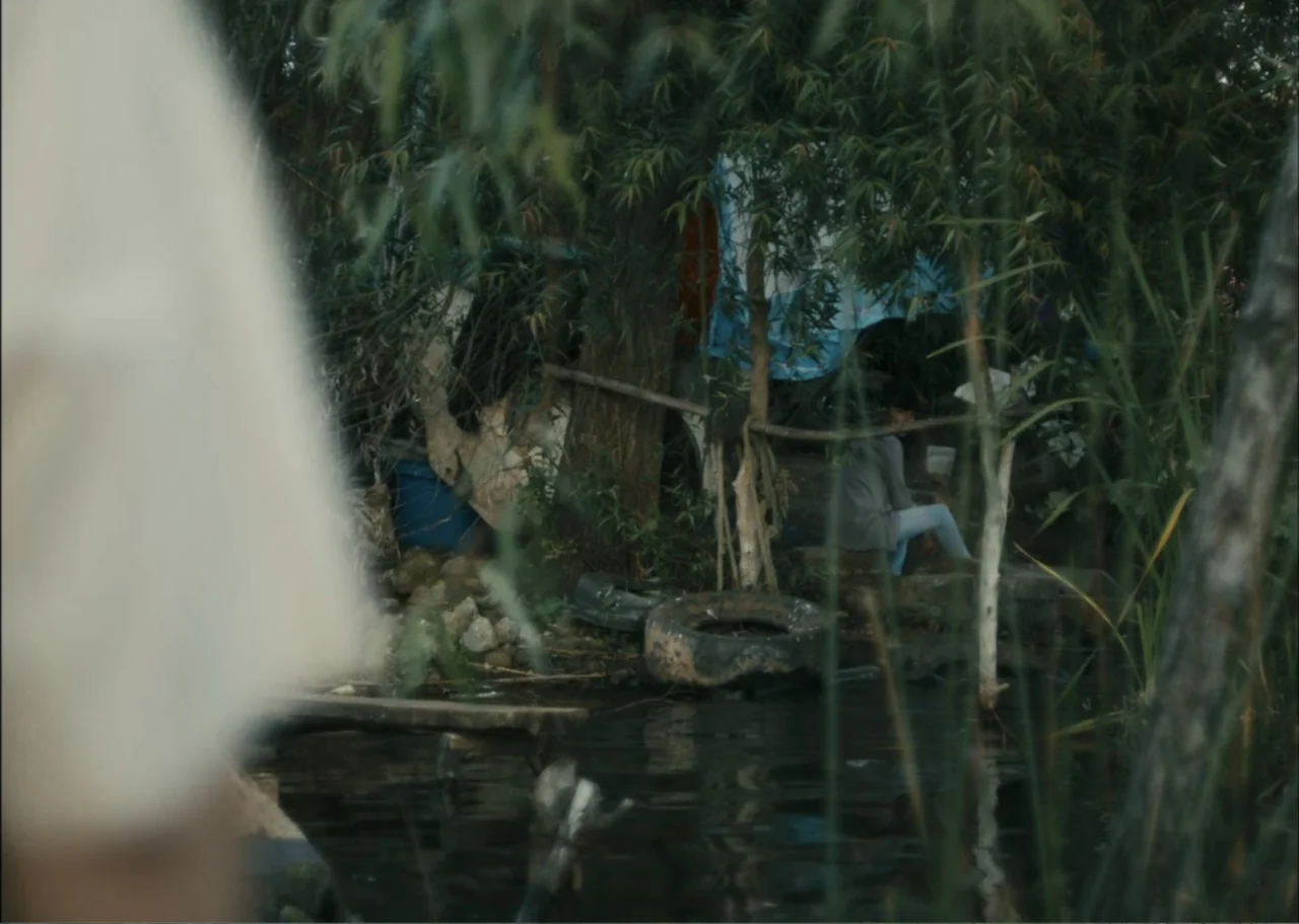 A Fisherman's Tale (1976) movie still: wide — A riverbank area filled with tall reeds and overgrown trees by a dark stream. A young woman sits alone…