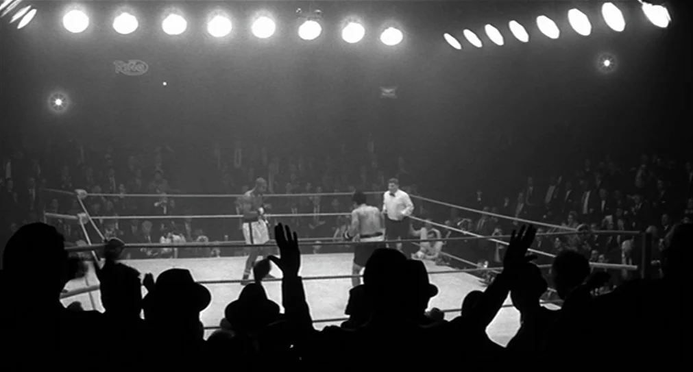 The Hurricane (1999) movie still: wide — An indoor boxing match is illuminated by a row of intense overhead arena lights. Two athletes move across…