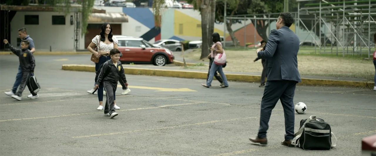 Divine Intervention (2026) movie still: fullbody — A paved lot marked with parking lines features a man facing a young boy. Other pedestrians walk…