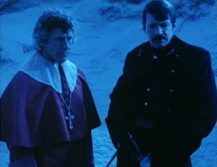The Tempest (1979) movie still: medium — A man in clerical robes and a man in a military coat stand together. They are situated in a sandy landscape…