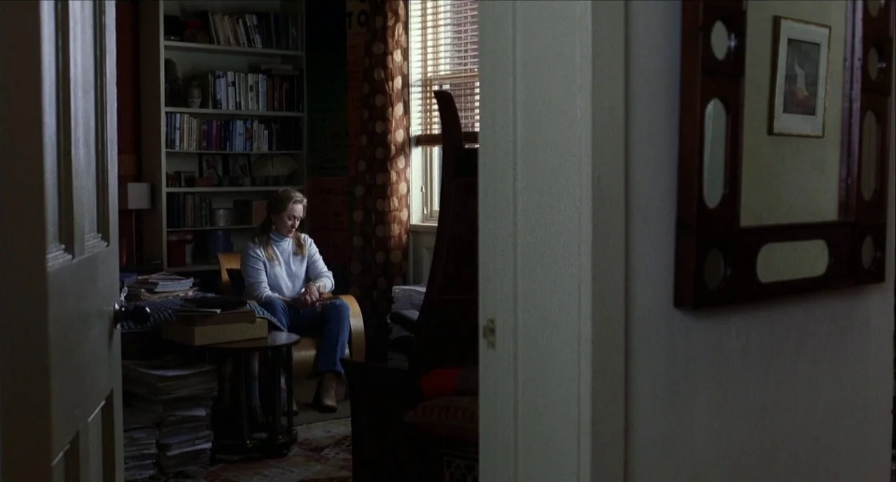 The Hours (2002) movie still: medium — A cluttered room features a tall bookshelf filled with various books against the wall. A woman sits in a…