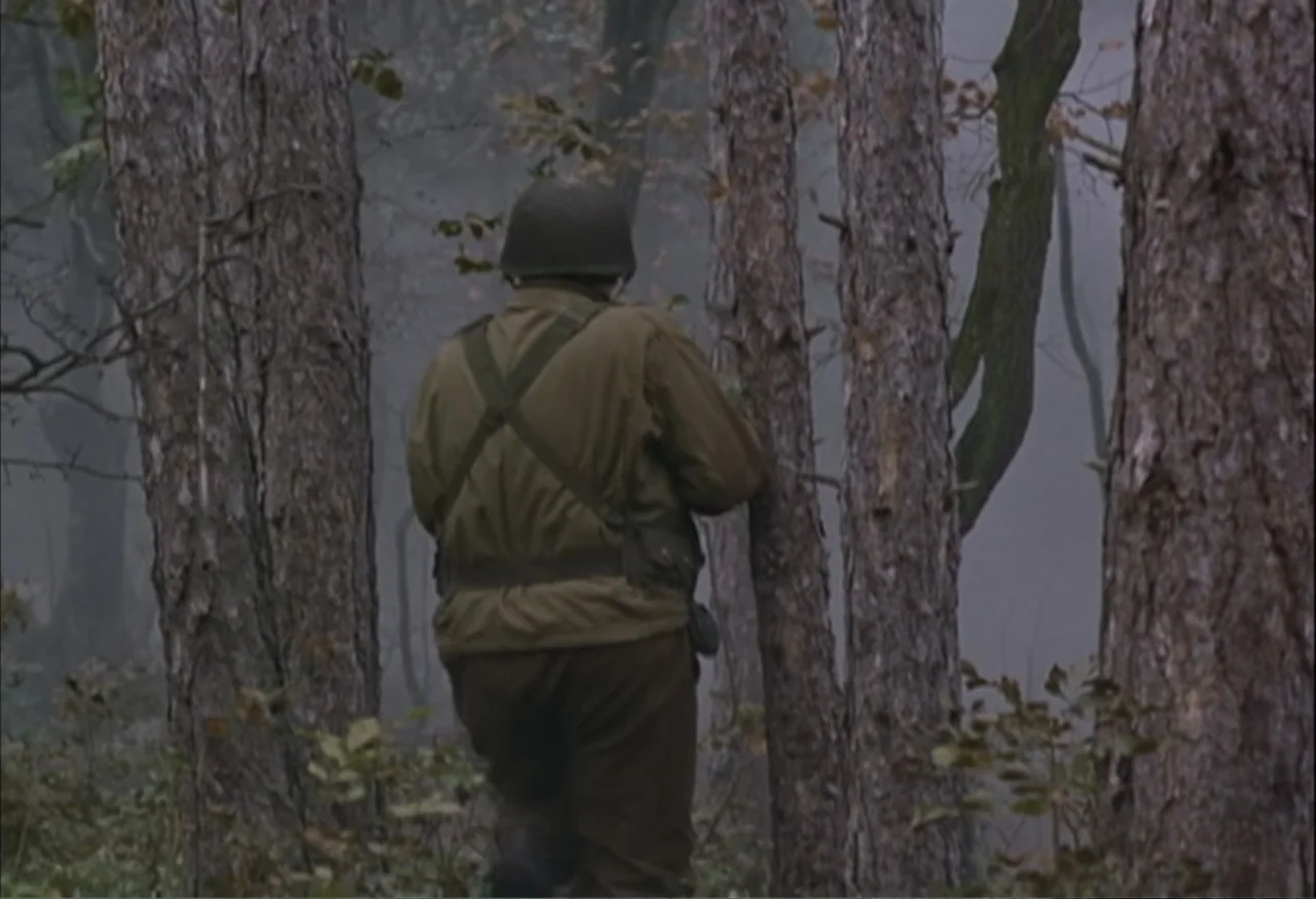 When Trumpets Fade (1998) movie still: medium — A soldier in military uniform walks through a dense woodland area during the daytime. The…